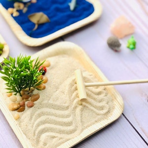 May include: A wooden tray with a sand garden design. The tray has a small rake and a few decorative stones. The sand is white and has a wavy pattern.