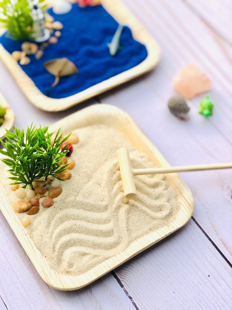 May include: A wooden tray with a sand garden design. The tray has a small rake and a few decorative stones. The sand is white and has a wavy pattern.