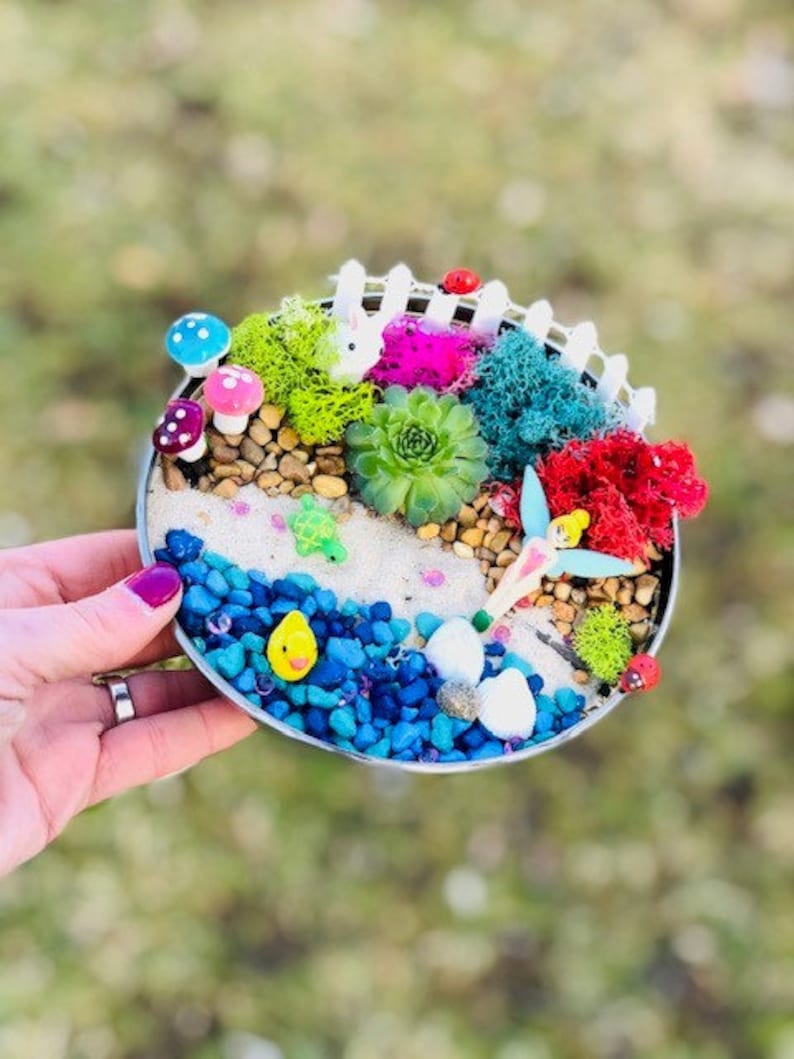 May include: A miniature fairy garden scene in a round metal container. The garden features a white picket fence, a white rabbit, a pink flower, green moss, blue and white stones, a yellow duck, and a white butterfly.