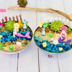 May include: Two miniature mermaid fairy gardens with a pink and a purple mermaid figurine, a small lighthouse, a white picket fence, and a succulent plant in each. The gardens are in a round, silver bowl with blue and brown rocks, sand, and a small turtle figurine.