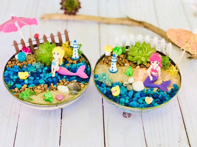 May include: Two miniature mermaid fairy gardens with a pink and a purple mermaid figurine, a small lighthouse, a white picket fence, and a succulent plant in each. The gardens are in a round, silver bowl with blue and brown rocks, sand, and a small turtle figurine.