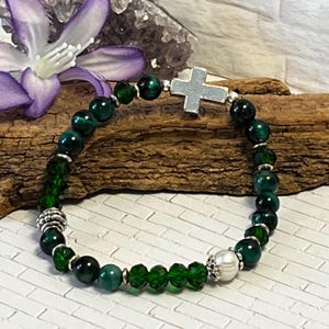 May include: A beaded bracelet featuring dark green and clear beads, with a silver cross charm. The bracelet includes a mix of round and faceted beads, and a white pearl accent. The bracelet is displayed on a white brick surface.