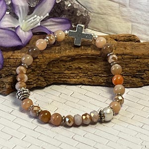 May include: A beaded bracelet featuring a silver cross charm and a mix of peach, brown, and orange-colored beads. The bracelet includes a pearl bead and decorative silver accents. The beads are strung on an elastic band.