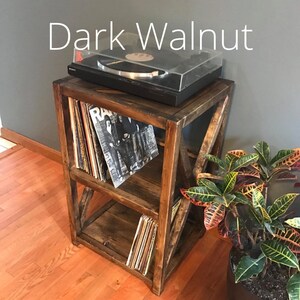 Pu&ograve; includere: Un supporto per giradischi in legno di noce scuro con due ripiani per riporre i dischi in vinile. Il supporto ha una finitura rustica e usurata e presenta un design a croce sul davanti.