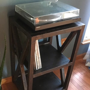 Pu&ograve; includere: Un supporto per giradischi in legno marrone scuro con due ripiani. Il supporto ha un design a X unico ed &egrave; etichettato "Ebony". Un giradischi nero con un coperchio trasparente si trova sul ripiano superiore. Uno stack di dischi in vinile si trova sul ripiano inferiore.