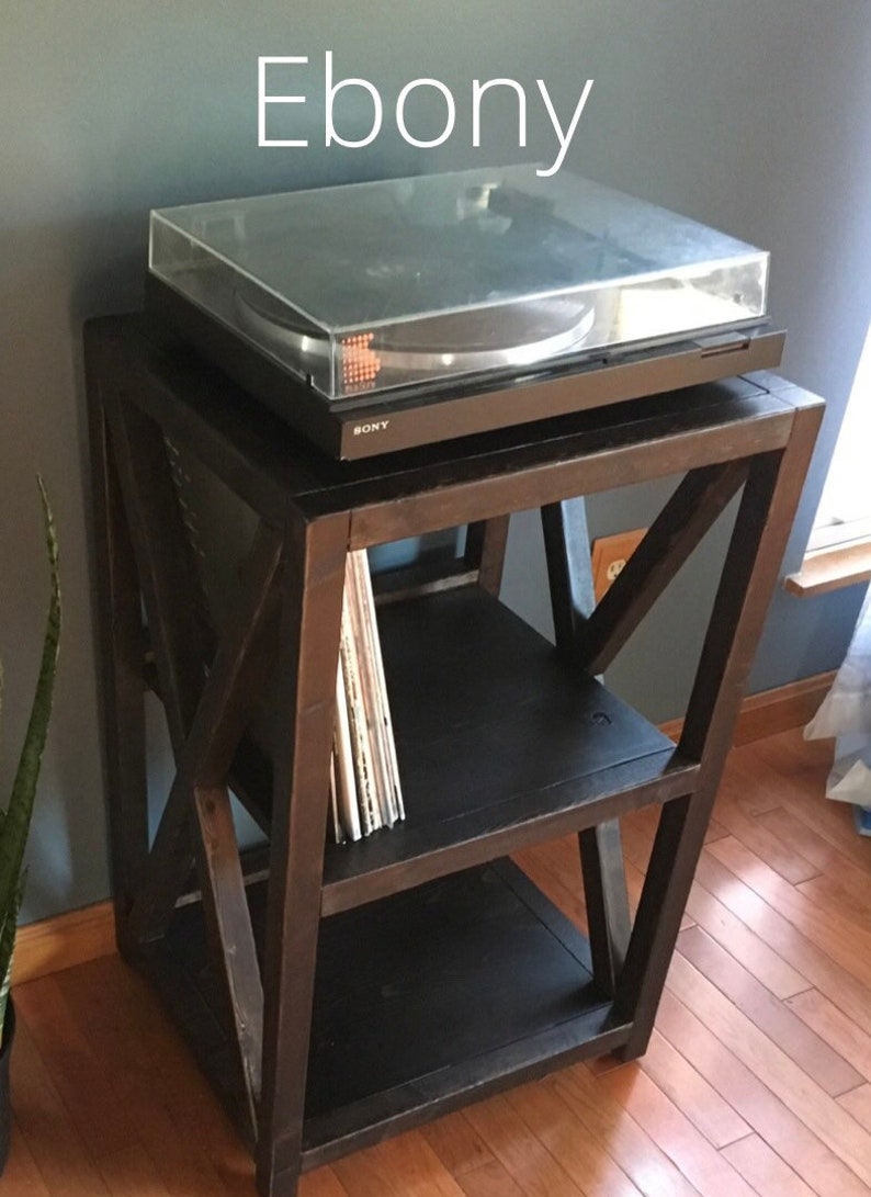 Pu&ograve; includere: Un supporto per giradischi in legno marrone scuro con due ripiani. Il supporto ha un design a X unico ed &egrave; etichettato "Ebony". Un giradischi nero con un coperchio trasparente si trova sul ripiano superiore. Uno stack di dischi in vinile si trova sul ripiano inferiore.