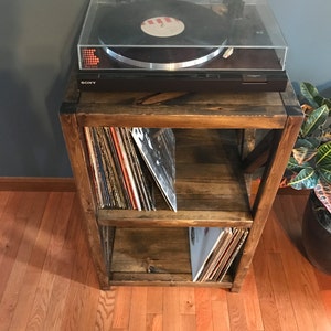 Pu&ograve; includere: Un supporto per giradischi in legno con due ripiani, che contiene una collezione di dischi in vinile. Un giradischi nero con un coperchio trasparente &egrave; posto sopra il supporto. Il supporto &egrave; in legno rustico con una macchia marrone scuro.