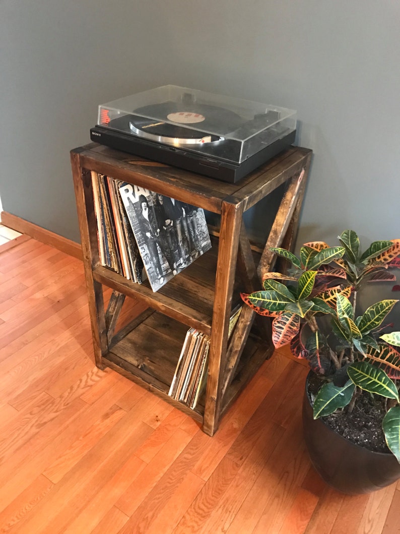 Pu&ograve; includere: Un supporto per giradischi in legno con due ripiani, che contiene dischi in vinile. Il supporto ha una finitura marrone rustica e un design a croce sul davanti. Un giradischi nero con un coperchio trasparente &egrave; posto sopra il supporto.