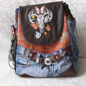 Op de afbeelding: Een unieke rugzak gemaakt van hergebruikte denim en leer. De flap heeft een geborduurd ontwerp van een schedel, geweien en vlinders. De tas heeft een bruine leren riem en een zilveren gesp, met een geweven stoffen paneel.
