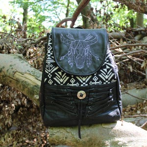 May include: A black backpack featuring a white embroidered bull skull design on the top flap. The bag includes a black and white geometric patterned fabric panel, black leather-like material, and a decorative lace-up front with a silver button.