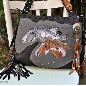 Könnte beinhalten: Grau-schwarze Leder-Umhängetasche mit gestickten Pferdeköpfen. Die Tasche hat einen silbernen Concho, Lederfransen, Federn und einen verstellbaren Riemen. Die Tasche steht auf einem weißen Stuhl.
