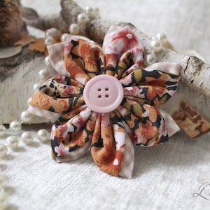 May include: A fabric flower brooch with a pink button in the center. The flower is made of multiple layers of fabric in shades of orange, brown, and white with a floral pattern. The brooch is surrounded by a string of white pearls.