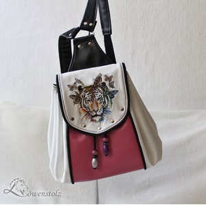 Op de afbeelding: Een witte en roze rugzak met een zwarte leren flap en een geborduurde tijger op de voorkant. De tijger wordt omringd door vlinders. De rugzak heeft een zwarte band en een zwarte rits.
