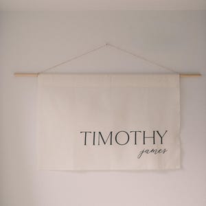 May include: A cream-colored banner with the name "TIMOTHY" in a bold, black font, and "james" in a smaller, cursive font. The banner is suspended from a wooden dowel rod with a string. The banner hangs against a white wall.