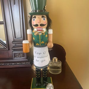 May include: A wooden nutcracker figurine in a green top hat and vest, holding a beer stein. The nutcracker has a painted face with a mustache and is wearing a white apron with the text "Top O' the mornin' to ya". The base is green and the boots are black.