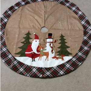 May include: A brown and tan Christmas tree skirt with a plaid border. The skirt features a snowman, Santa Claus, and two reindeer in a snowy scene.