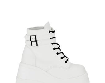 May include: White platform boots with black laces and a buckle closure. The boots have a chunky sole and a short ankle height. The boots are made of a smooth, solid white material.