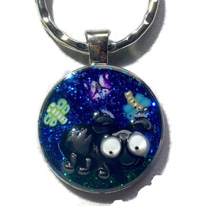 May include: A black and white cartoon creature with big eyes is surrounded by glitter and colourful butterflies in a blue resin keychain.