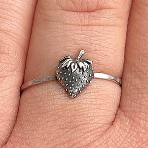 May include: A silver ring featuring a strawberry design. The strawberry has a textured surface and a small stem. The ring is worn on a finger, showcasing the delicate details of the jewelry.