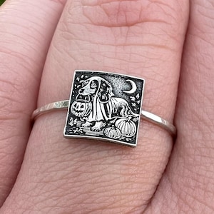 Może przedstawiać: Srebrny pierścionek z kwadratową zawieszką z grawerunkiem przedstawiającym jamnika w kostiumie na Halloween, dynie i półksiężyc. Pierścionek ma cienką obrączkę i jest noszony na palcu.