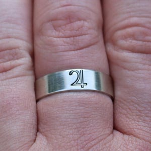 Può includere: Un anello d'argento con fascia piatta, con un simbolo nero di Giove. L'anello è indossato su un dito, con uno sfondo verde sfocato.