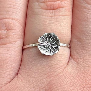 May include: A silver ring featuring a detailed, textured leaf design. The leaf is the focal point, with intricate veining. The ring band is thin and delicate, adding to the ring's elegant and natural aesthetic. A nature-inspired piece of jewelry.