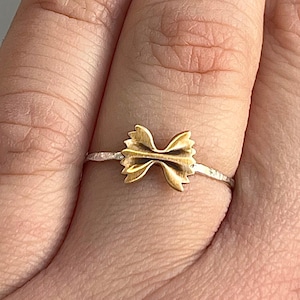 Pode incluir: Um anel dourado com um design de laço. O anel está montado numa banda fina prateada. O laço é o ponto focal, com curvas detalhadas e um acabamento polido. O anel é mostrado no dedo de uma pessoa.