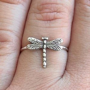 May include: A silver dragonfly ring. The ring features a detailed dragonfly design with outstretched wings and a segmented body. The ring is worn on a finger, showcasing the delicate craftsmanship and nature-inspired theme.