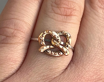 May include: A gold-colored pretzel ring. The ring features a detailed pretzel design, with a textured surface. The ring is set on a thin, silver-colored band. The ring is shown on a finger.