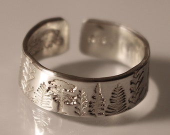 Puede incluir: Anillo ajustable plateado con una escena de bosque grabada alrededor de la banda. El diseño incluye árboles y follaje. El anillo está abierto en la parte superior para ajustar la talla. El anillo se muestra sobre una superficie clara.
