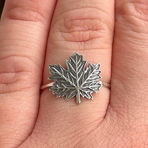 May include: A silver ring featuring a detailed maple leaf design. The ring's band is thin, and the leaf is the focal point, showcasing intricate veining and texture. The ring is a nature-inspired accessory.
