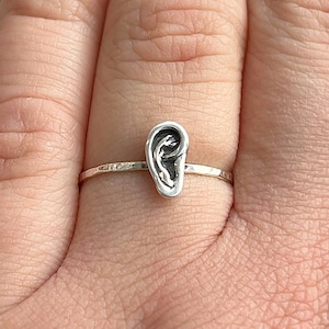 May include: A silver ring featuring a small, detailed ear design. The ear is a three-dimensional sculpture with intricate details. The ring is shown on a person's finger, highlighting the delicate craftsmanship and unique style of the jewelry.