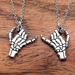 May include: Two silver necklaces with skeleton hand pendants. The hands are facing each other and making a heart shape.