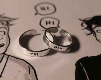 May include: Two silver rings with the word "HI" and a heart symbol engraved on them. The rings are on a white surface with a cartoon drawing of two people in the background.