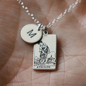 Puede incluir: Collar de plata con un dije de la carta del tarot Fuerza y un dije inicial personalizado. El dije inicial es un círculo con la letra "M" grabada en él.