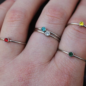 May include: Five simple silver rings with small colorful gemstones. The rings are stacked on a person's fingers. The gemstones are red, blue, clear, green, and yellow.