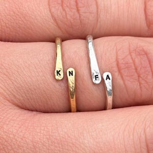Puede incluir: Dos anillos abiertos, uno dorado y otro plateado, con textura martillada. El anillo dorado está estampado con las letras K y N, mientras que el anillo plateado lleva las letras F y A en sus respectivos extremos planos.