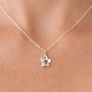 Puede incluir: Un collar de plata con un colgante de flor. El colgante tiene una pequeña gema roja en el centro. El collar está suspendido de una fina cadena. Una joya.