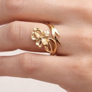 May include: A gold-colored ring featuring a detailed floral design. The ring's focal point is a sculpted flower, with delicate petals and a stem that wraps around the finger. The ring is made of a shiny, metallic material.