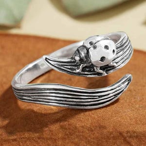 May include: A silver-toned ring featuring a ladybug design. The ring wraps around the finger with a leaf-like detail. The ladybug has a textured body with visible spots. The ring is set against a brown surface.