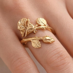 May include: A gold-coloured ring with a rose design. The ring wraps around the finger with a rose bud and leaves. The rose and leaves have textured surfaces. The ring is a piece of jewellery.