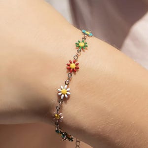 Könnte beinhalten: Ein silbernes Armband mit bunten Gänseblümchen-Anhängern. Die Gänseblümchen-Anhänger haben gelbe Zentren und Blütenblätter in verschiedenen Farben, darunter Weiß, Rot, Grün und Blau. Das Armband wird am Handgelenk getragen.
