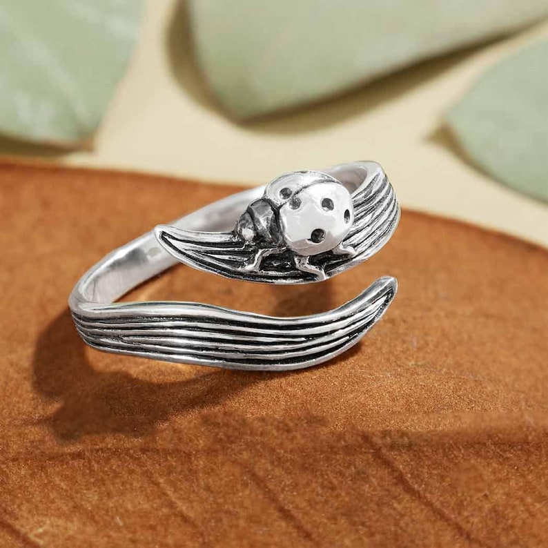 May include: A silver-toned ring featuring a ladybug design. The ring wraps around the finger with a leaf-like detail. The ladybug has a textured body with visible spots. The ring is set against a brown surface.