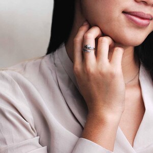 May include: A silver-toned ring with a floral design and a small, clear gemstone. The ring is wrapped around the finger, with the floral design on the top. The ring is worn on the ring finger.