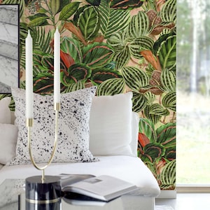 May include: A white couch with two white throw pillows and a black and white patterned throw pillow. A gold candle holder with two white candles is on a glass coffee table in front of the couch. The wall behind the couch is covered in a green and red tropical leaf wallpaper.