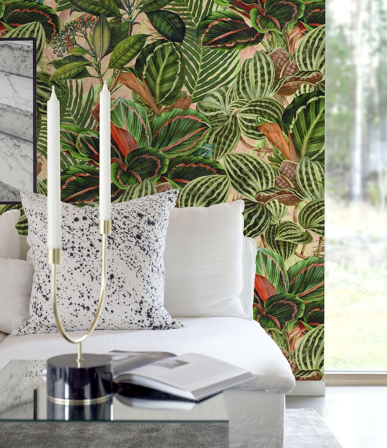 May include: A white couch with two white throw pillows and a black and white patterned throw pillow. A gold candle holder with two white candles is on a glass coffee table in front of the couch. The wall behind the couch is covered in a green and red tropical leaf wallpaper.
