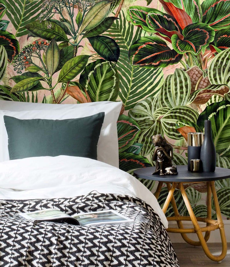 May include: A white bed with a black and white patterned blanket and a green pillow. The bed is in front of a wall with a tropical leaf wallpaper. A small wooden side table with a black top holds a small statue and two gold candle holders.