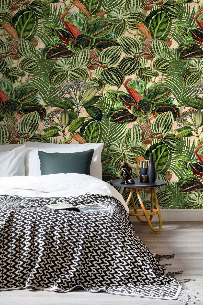 May include: A white bed with a black and white patterned throw blanket. The bed is in front of a wall with a tropical leaf pattern wallpaper. A small side table with a black top and a woven base sits next to the bed.