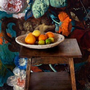 Op de afbeelding: Een geweven mand gevuld met sinaasappelen, citroenen en limoenen staat op een donkerhouten tafel met twee planken. De tafel staat voor een schilderij met bloemen en twee engelen.