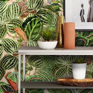 May include: A white metal shelf unit with two shelves, featuring a tropical leaf patterned wallpaper backdrop. The top shelf has a white ceramic bowl with a small green plant, a gold metal vase, and a terracotta vase. The bottom shelf has a white ceramic pot with a small green plant and a wooden serving tray.